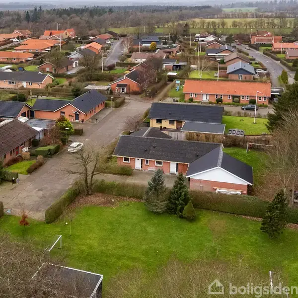 Et boligområde med rødstenshuse omgivet af grønne haver og træer. Mindre veje løber mellem husene. Området ligger i en moderne, tæt boligbebyggelse på landet med åbne marker i baggrunden.