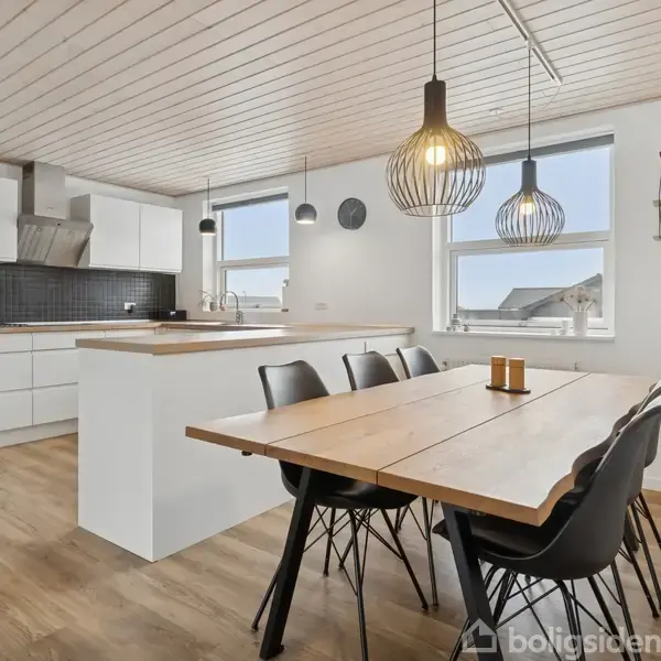Spisebord med seks sorte stole står i et lyst køkken-alrum. To hængelamper hænger over bordet. Køkkenet har hvide skabe og en mørk væg. Store vinduer giver naturligt lys.