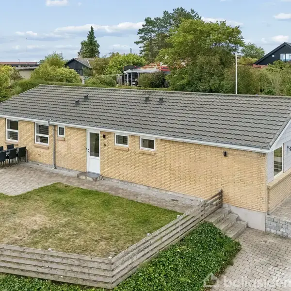 En étplans villa med gult murværk og sadeltag står på en flisebelagt terrasse. Et havebord med stole er placeret udenfor. Omgivet af rækkehuse og grønne arealer.