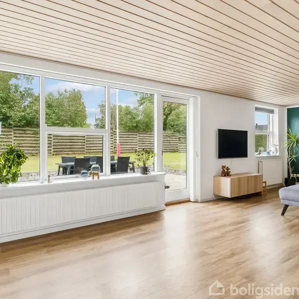 Stue med trægulv og lys træloft. Sofa står langs højre væg, mens store vinduer og havedør på venstre væg giver udsigt til en grøn have. TV monteret på væggen.