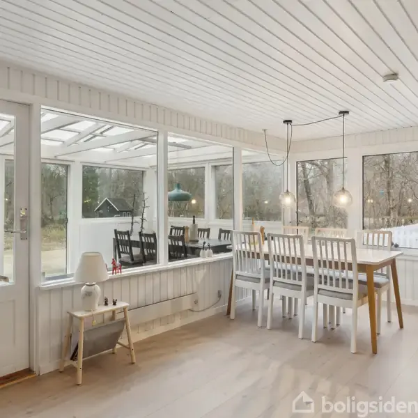 Spisebord omgivet af hvide stole står i et lyst rum med store vinduer, der åbner op til en terrasse og udsigt til natur udenfor. To lamper hænger over bordet.