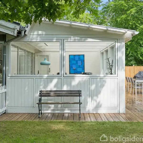 Brunt læderlænestol står i lyse stue, mens et langt spisebord med hvide stole ses ved vinduerne. Væggene er hvide og rummer billeder og dekorationer, med lysindfald gennem store vinduer.