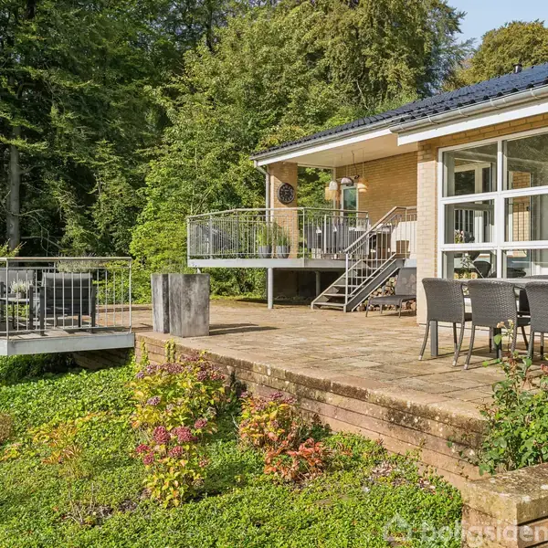 Et moderne hus har store vinduer og en terrasse med udendørs møbler. Terrassen strækker sig ud over en grøn have, omgivet af høje træer i et skovområde.