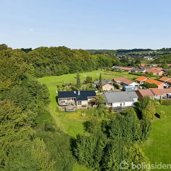 Et hus med sort tag ligger i en grøn landskabskontekst, omgivet af tæt bevoksning og træer, med en lille boligområde i baggrunden.
