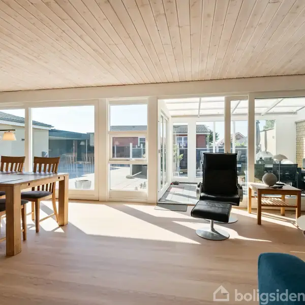 Stue med spisebord og stole i træ til venstre, og sort lænestol foran vinduer med udsigt til terrasse. Sollys strømmer ind, skaber en lys og åben atmosfære.