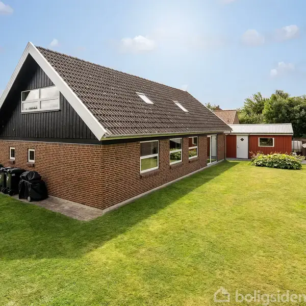 Rødstenshus med mørkt sadeltag, omgivet af en velplejet grøn have. Flere vinduer pryder husets side, og en skraldespand står mod siden af huset. Klart solrigt miljø.