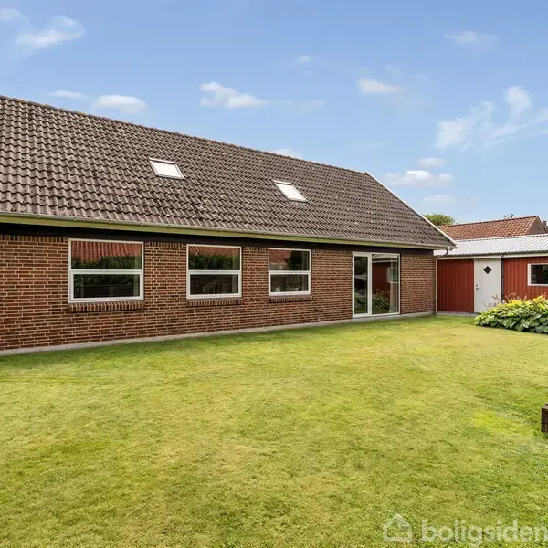 Et rødt murstenshus med skråt tag og fire vinduer står i en velplejet have med græsplæne. I baggrunden ses en rød skurbygning under en klar blå himmel.