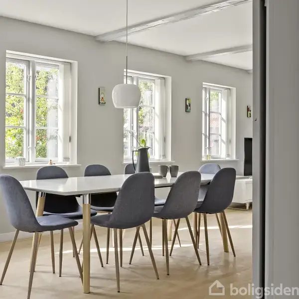 Et spisebord med seks blå stole står i et lyst rum. En moderne lampe hænger over bordet. Tre store vinduer tillader naturligt lys at strømme ind.