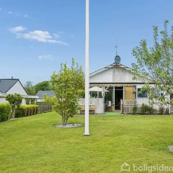 Et lille hus med terrasse står centralt i en velholdt have med græsplæne, træer og flagstang. Flere tilstødende huse og blå himmel ses i baggrunden.