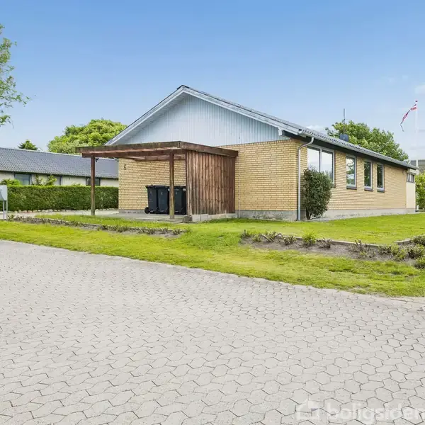 Hus af gul mursten med sort tag, udenfor står affaldsspande under et halvtag. Græsplæne omringer huset, belægningssten danner indkørsel. Flagstang ses i baggrunden.