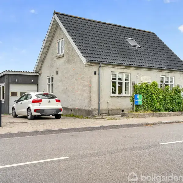 Huset står ved en vej, med en bil parkeret i indkørslen. Det har en lys facade og sort tag. En grøn hæk løber langs siden tæt ved vejen.