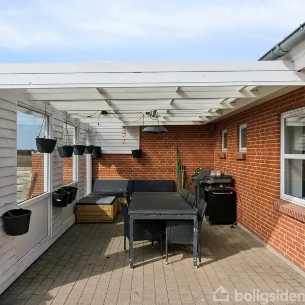 En overdækket terrasse med et sort bord og stole samt en grill under et hvidt trætag. Omkranset af røde murstensvægge med hængende sorte potter og vinduer.