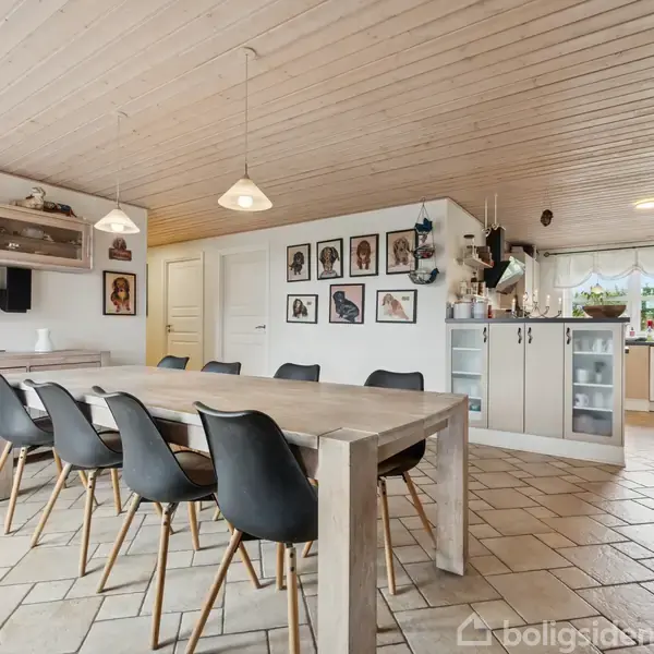 Langt spisebord med sorte stole står i et åbent køkken-alrum. Trægulve, lys træloft og hvide skabe bidrager til en lys atmosfære. Dekorationer og kunst pryder væggene.