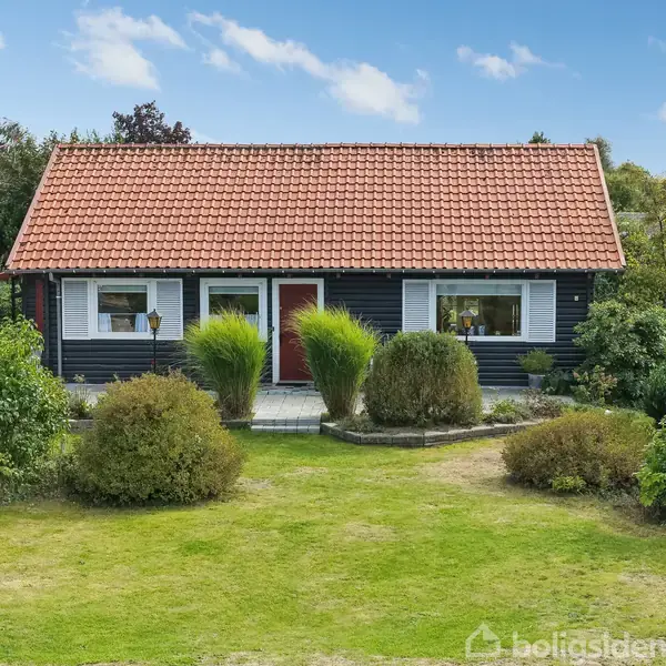 Hus står i en frodig have, har rødt tegltag og sort facade. Store buske omgiver huset, og græsplæne strækker sig foran. Klart solskin oplyser området.