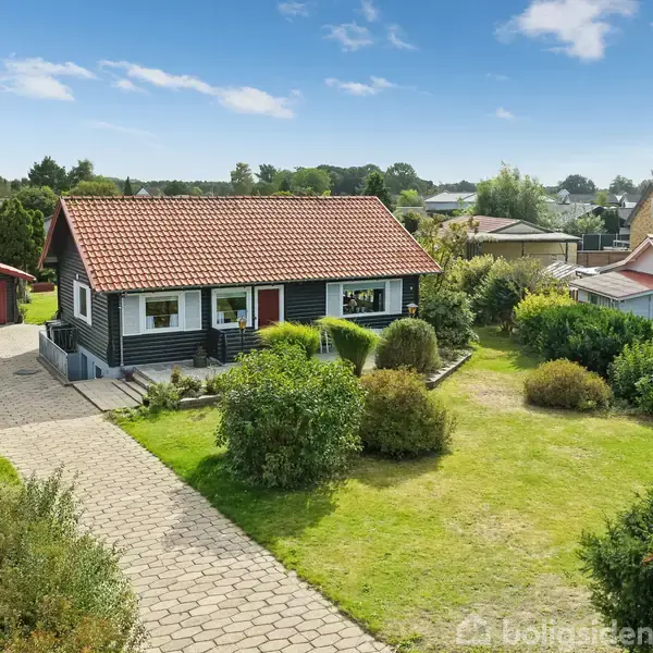 Et lille, sortmalet hus med rødt tegltag står i en velplejet have med grønne buske og en stenbelagt sti. Det er omgivet af andre huse i en rolig, landlig bebyggelse.