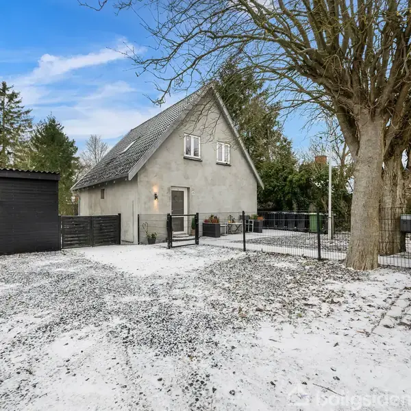 En grå murstenshus står roligt ved siden af en stenet indkørsel med sort skur i en snedækket have omkranset af høje træer under en klar blå himmel.