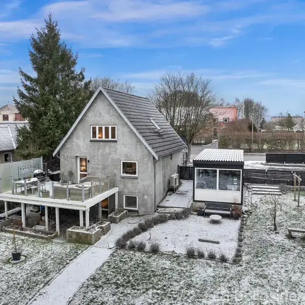 Et gråt hus med gavltag står i en snedækket have, hvor der er en stor træterrasse ovenpå en carport. Baggrunden viser andre huse og træer under en blå himmel.