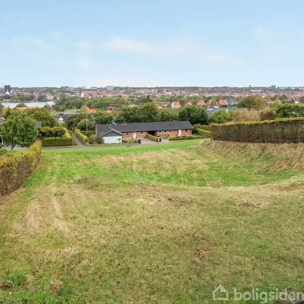 Græsplæne ligger stille mellem to hække i et boligområde med udsigt over en by i baggrunden. Huse omkranser området, skaber en følelse af forstadsmiljø.