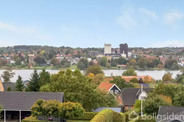 Huse med mørke tage står blandt træer og buske. I baggrunden ses en sø og en by med bygninger og træer under en blå himmel.