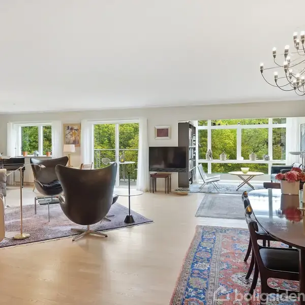 En elegant stue med moderne møbler, herunder en skulpturel stol og bord, foran store vinduer. Rummet er lysfyldt, med en lysekrone og udsigt til frodige, grønne omgivelser.