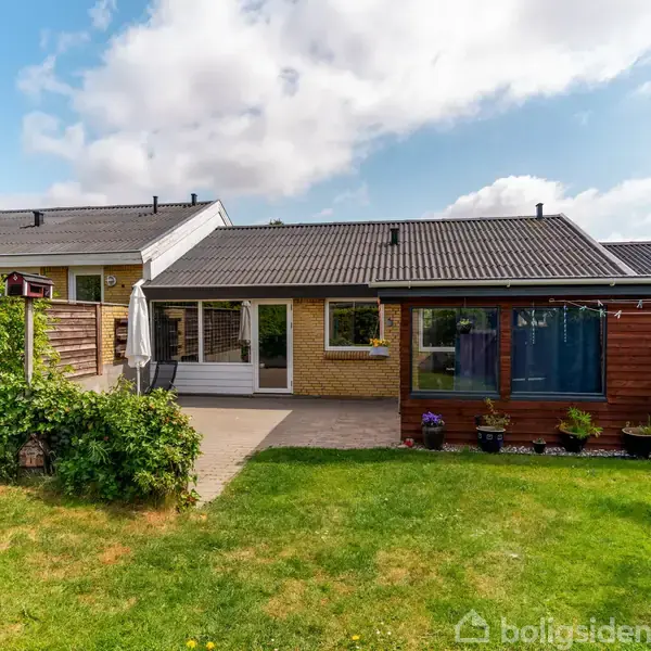 Hus med fladt tag og store vinduer står i en frodig have. Ved siden af er en terrasse med parasol. Skyfri himmel og solrig dag.