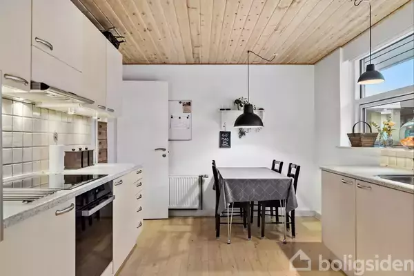 Et lyst køkken med trægulv og træloft, hvor et spisebord med dug og fire stole står ved en hvid væg. Ved siden af er der et vindue, og overskabene er lyse. Der hænger lamper over bordet.