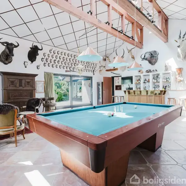 Et billardbord står centralt i et lyst rum med trofæer på væggene, herunder dyrehoveder. Rummet har store vinduer, en pejs, og en bar i baggrunden.