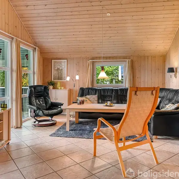 Stue med trævægge og loft. En lænestol og sofa står omkring et sofabord på et tæppe. Fladskærms-TV til venstre, brændeovn til højre. Store vinduer giver udsigt.