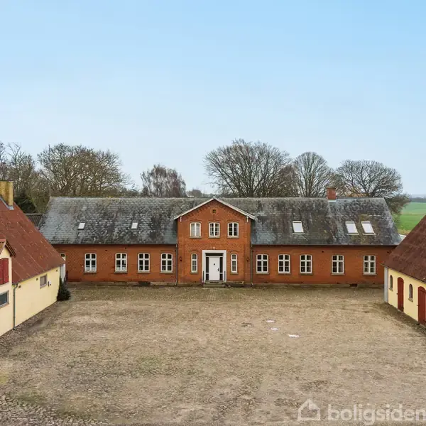 En historisk trelænget gård med røde mursten udgør midten, omkranset af to gule bygninger med røde tage. Gårdspladsen er grusbelagt, og bagved strækker sig et grønt landskab.