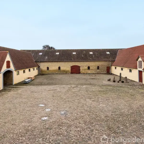 En gårdsplads omgivet af flere længers bygninger med røde tage og døre. En åben port og stenbelagt underlag giver et traditionelt gårdsmiljø med åbent himmelrum.