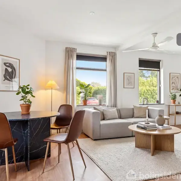En stue med en lys grå sofa og rundt sofabord står centralt. Store vinduer slipper lys ind, omgivet af planter og minimalistisk kunst på væggene. "Visualisering" er skrevet øverst til højre.