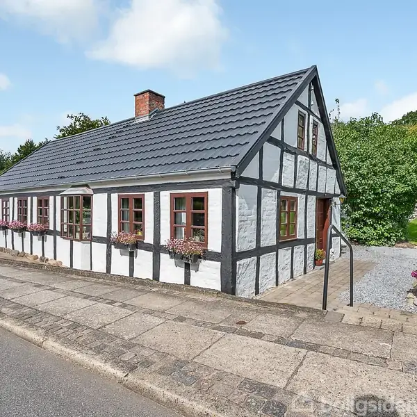 Hus med hvid facade, sort bindingsværk og røde vinduer står stille på en brolagt vej omgivet af grønne buske og træer under en blå himmel med lette skyer.