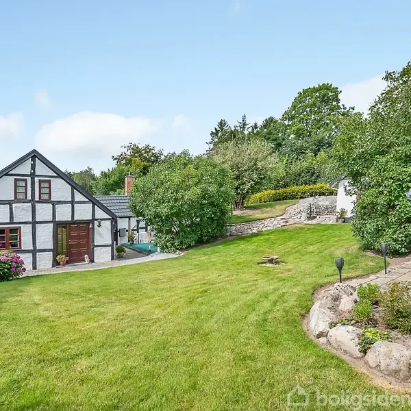 Et bindingsværkshus står i en velplejet have med græsplæne og buske. Til højre ses en gyngesofa, og der er træer i baggrunden under en blå himmel.