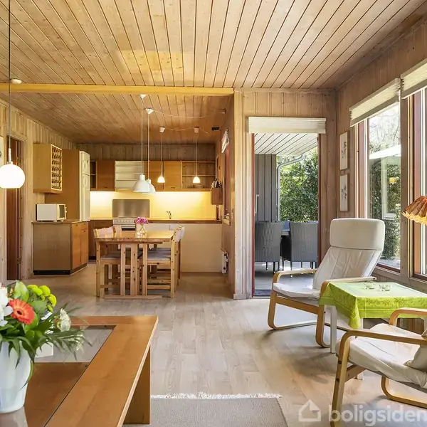Stue med trævægge og loft. Hvid lænestol ved stort vindue, nær grøn lampe. Spisebord med stole centralt. Køkken i baggrunden. Blomsterdekoration på sofabord.