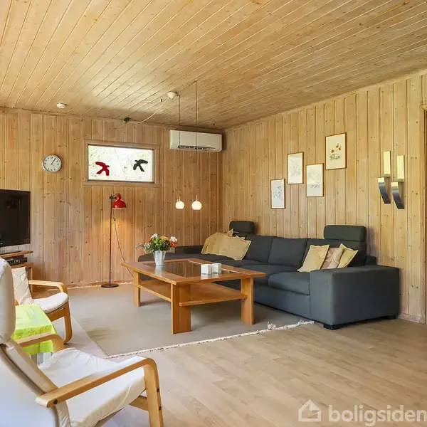 Stuen har træbeklædte vægge og loft, en grå sofa, træbord, og lænestol. Et fjernsyn står på en reol. Væggene har billeder, og der hænger lamper.