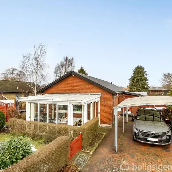 En murstenshus med vinduesveranda, omgivet af en tætklippet hæk, står i en have med snepletter. En carport med to biler til højre, mens et dansk flag vajer.