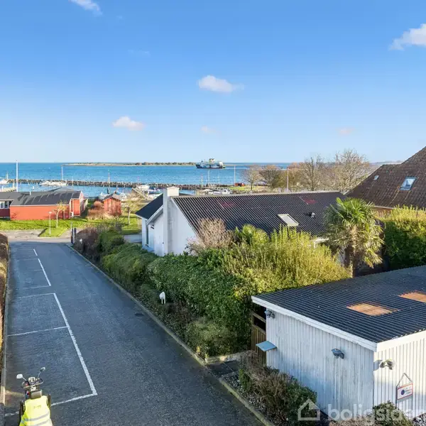 En kystnær gade med huse langs vejen, der fører mod en havn fyldt med både. Baggrunden viser en blå himmel og horisonten med hav.