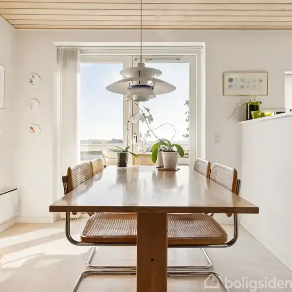 Et spisebord med seks stole står i et lyst rum. Bordet er dækket med planter og lamper hænger over det. Store vinduer giver udsigt til det fri og indretningen er minimalistisk.