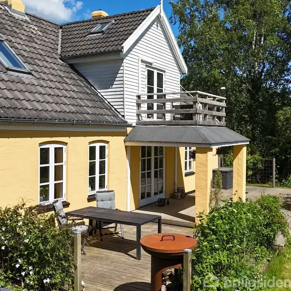 Et gult hus med sorte tagsten står i en have. En træterrasse med borde og stole strækker sig fra huset. Grønne planter omgiver området, og solen skinner klart.