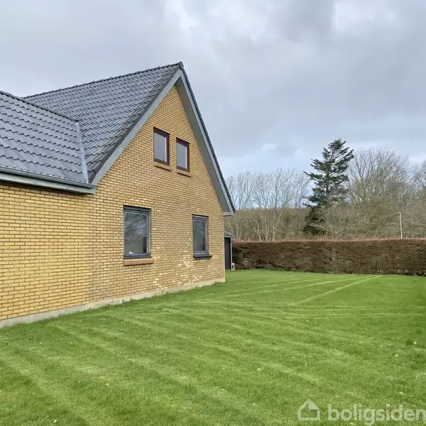 Et gult murstenshus med blågråt tag står i en velplejet have. Huset har flere vinduer, og i baggrunden ses en hæk og nogle træer.
