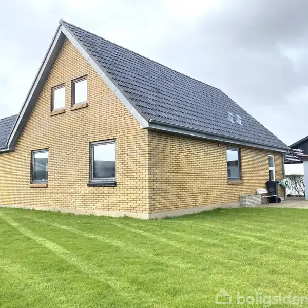 Et gult murstenshus står på en velholdt græsplæne. Huset har en sort tagflade og flere vinduer. I baggrunden ses en lys garage eller skur ved siden af huset.