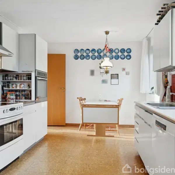 Et lyst køkken med hvide skabe og rødbrune fliser. Der er en ovn og en kogeplade til venstre. I baggrunden står et bord med to stole og tallerkener på væggen.