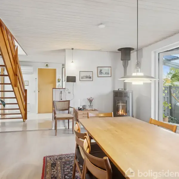 Spisebord med træstole står i spisestuen, mens en brændeovn brænder ved siden af. En trappe fører opad, og store vinduer vender ud mod en have. En dør ses i baggrunden.