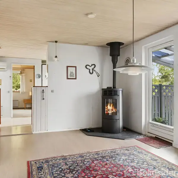 Garage i rød murstensbygning med brun dør og garageport. Murstensvæg til venstre og grøn busk på højre side i en flisebelagt indkørsel. Sollyst omgivelser med træer i baggrunden.