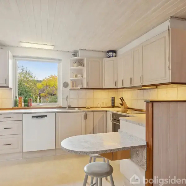 Et lyst køkken med hvide skabe og en opvaskemaskine. Der er et rundt bord med en stol. Et stort vindue giver udsigt til en have udenfor.