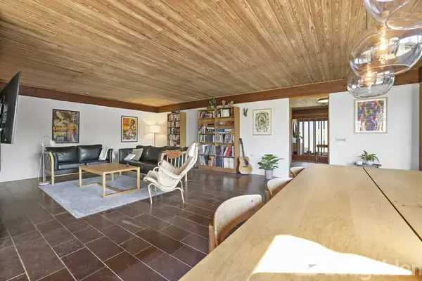 En stue indeholder sorte lædersofaer og træborde, flankeret af en bogreol med bøger og en guitar. Store vinduer giver lys ind, og væggene er dekoreret med kunstplakater.