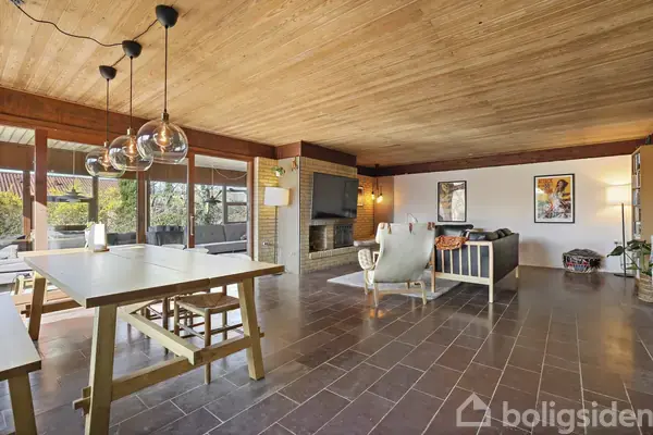 Et rummeligt stueområde med træloft og klinkegulv. Et spisebord med stole står til venstre, mens en sofa og lænestol er placeret omkring et TV på væggen og en pejs.