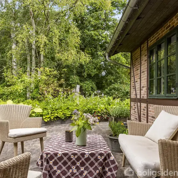 Terrassemøbler står på en stenbelagt terrasse. På bordet ses en blomstervase. Omgivet af frodig vegetation, er terrassen beliggende ved et murstenshus med sortmalede vinduesrammer.