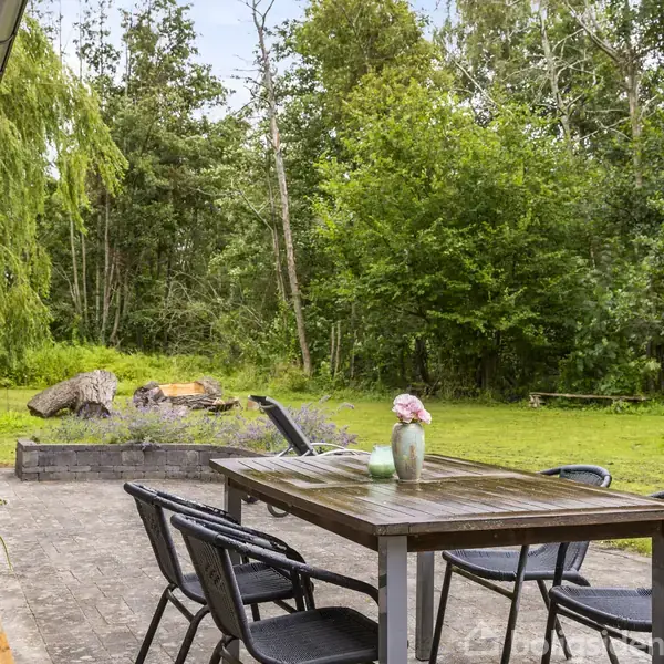 Træbord med stole står på en terrasse, omgivet af frodig have med græs og træer. På bordet står en grøn vase med blomster. Grill og bålplads ses i baggrunden.