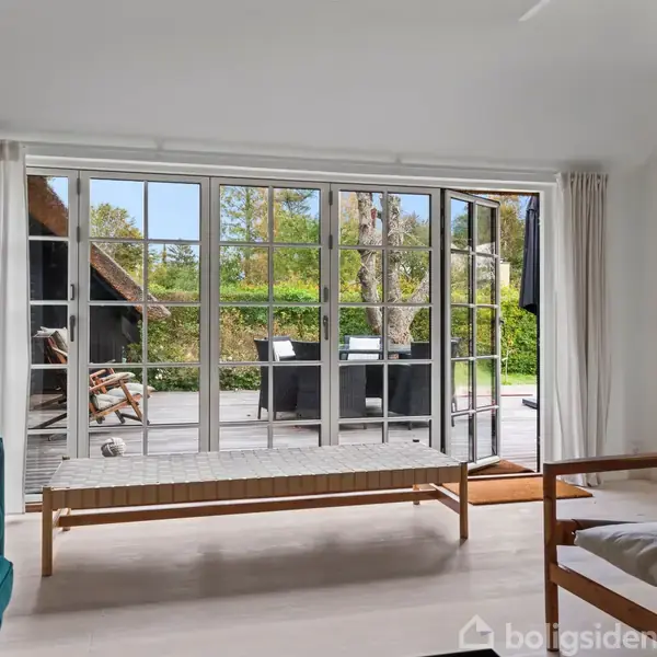 Daybed står i en lys stue med store vinduer, der giver udsigt til en terrasse med bord og grønne omgivelser. Foran er en grøn sofa og en lænestol.